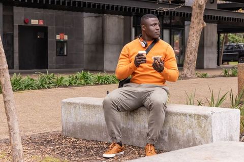 Commuter in orange sweater eating pastry and holding coffee on concrete bench