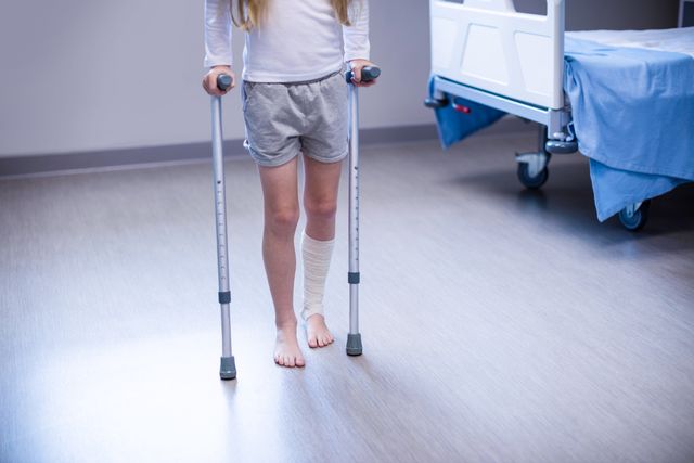 Young girl using crutches for support while walking in a hospital ward. Ideal for use in healthcare, medical recovery, pediatric care, and rehabilitation contexts. Can be used in articles, blogs, and educational materials about injury recovery, child healthcare, and hospital environments.