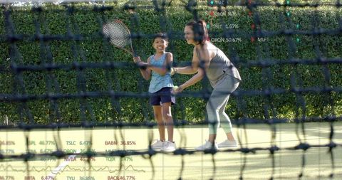 Coach guiding young girl during tennis lesson on sunlit court with green hedge backdrop