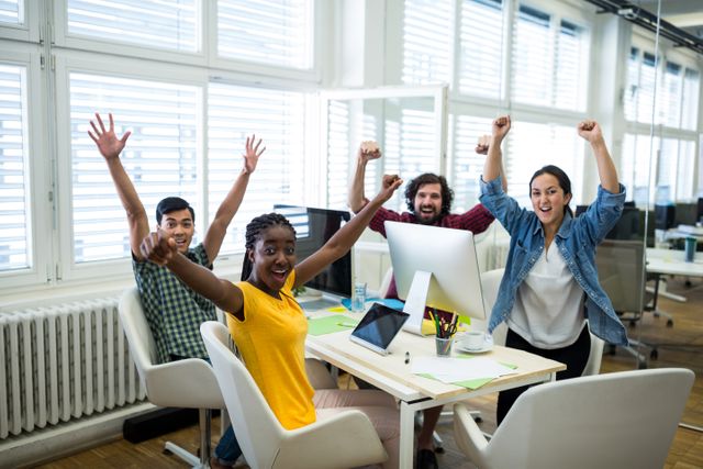 A group of diverse business executives celebrating success while sitting at an office desk. Their hands are raised in excitement, and they appear very happy. This image is ideal for use in marketing materials, corporate presentations, teamwork promotions, and business success stories.