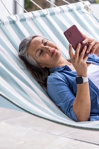Senior woman relaxing on striped hammock checking smartphone with smartwatch health alert