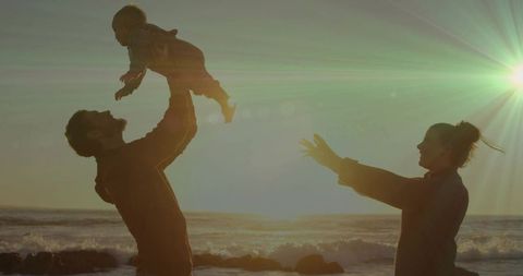 Father Lifting Baby Toward Mother on Beach at Sunset Silhouette