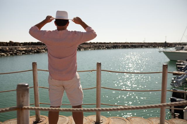 Rear view of Caucasian man, wearing a pink shirt and a hat, enjoying his time on a promenade by the sea on a sunny day, standing with arms in the air admiring the stunning view.