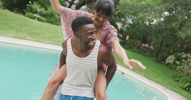 A young couple having fun near a swimming pool on a summer day. The man is giving a piggyback ride to the woman while both are laughing and enjoying their time. The lush greenery in the background adds to the natural, relaxed vibe. Useful for emphasizing fun, relaxation, outdoor activities, vacation, and relationships in marketing materials, social media posts, and lifestyle blogs.