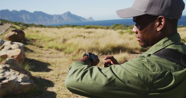 African American man in green jacket and baseball cap checking smartwatch in scenic location with mountains and sea in background. Suitable for travel, adventure, hiking, outdoor activities, technology and smartwatch-related themes.
