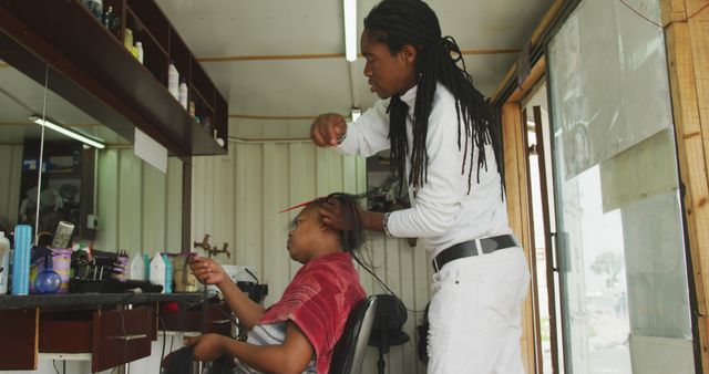 Man with long dreadlocks braiding another man's hair in a modern barber shop. Ideal for use in articles about hairdressing techniques, male grooming, hair care services, and professional barber services.