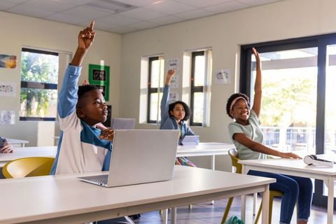 Elementary students raising hands and engaging with laptops in bright modern classroom