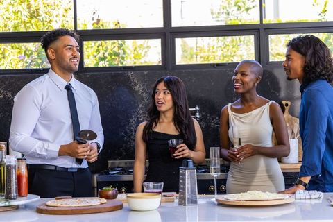 Multicultural friends preparing pizza and chatting in sunlit modern kitchen with drinks