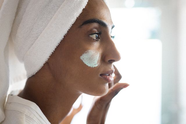 Close-up of biracial mid adult woman with towel wrapped on head applying facial mask. Unaltered, skin care, beauty, pampering, body care and wellbeing concept.
