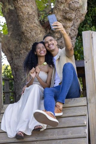 African american couple taking selfie on treehouse steps holding white rose smiling