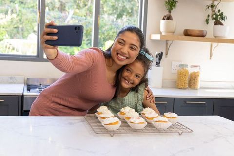 Indian mother and daughter taking selfie while baking cupcakes in sunlit modern kitchen