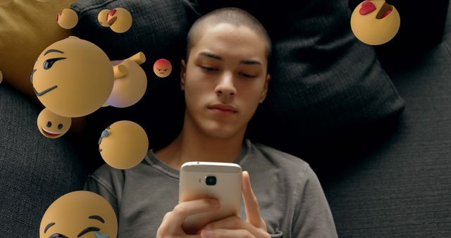 Teenage boy lying on a sofa scrolling through smartphone screen, various emojis float around him representing different emotions. Perfect for use in discussions about technology's effect on youth, social media engagement, digital communication trends, and modern lifestyles.