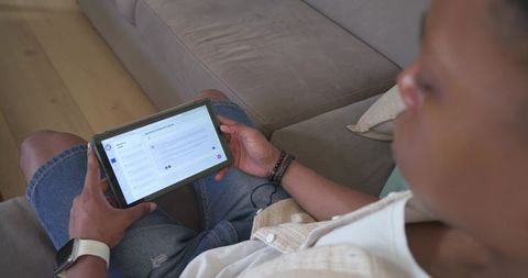 African american man lounging on sofa reading messages on tablet with earphones