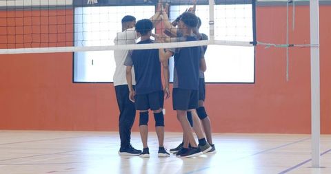 Diverse male volleyball team huddling with coach under net in gymnasium