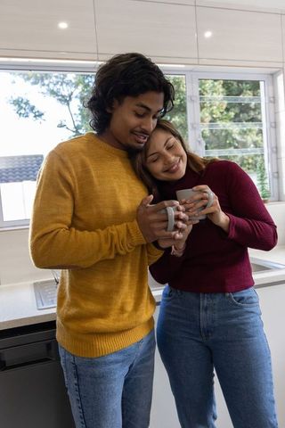 Indian couple sharing morning coffee in sunlit kitchen embracing cozy sweaters and warm connection