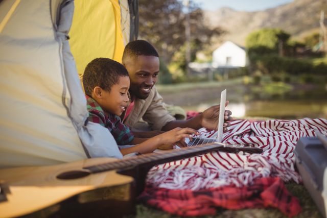 Father and son enjoying quality time together using a laptop inside a tent during a camping trip. Ideal for themes related to family bonding, outdoor activities, technology in nature, and leisure time. Can be used for advertisements, blog posts, or articles about family vacations, camping tips, or digital detox.
