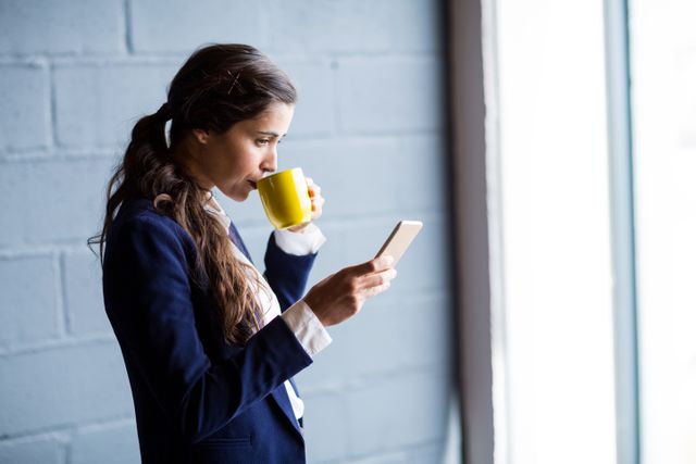 Young businesswoman in professional attire drinking coffee while using smartphone in modern office. Ideal for illustrating concepts of multitasking, corporate lifestyle, productivity, and modern work environments. Suitable for use in business blogs, corporate websites, and promotional materials.