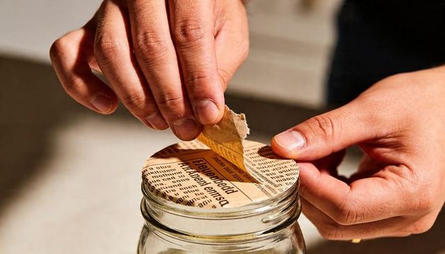 Peeling newspaper lid from glass jar with adult hands closeup for diy kitchen reuse