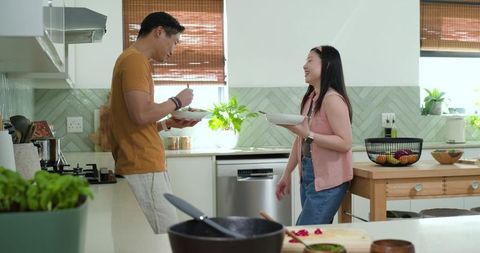 Asian couple preparing meal together in sunlit modern kitchen, laughing and sharing bowls