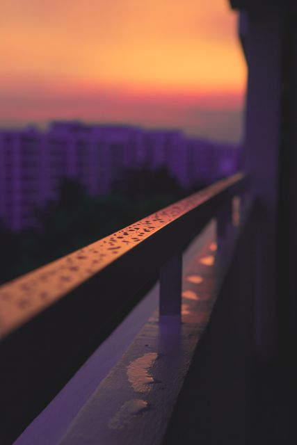 Sunset over urban setting with focus on wet balcony railing with raindrops, background showing out-of-focus buildings under purple and golden hues. Perfect for use in painting serenity, evening atmosphere in city, or concepts of rain and tranquility.