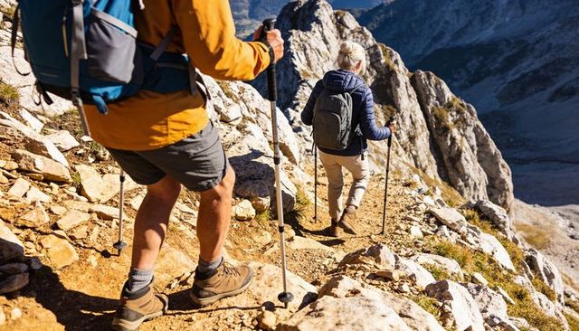 Two hikers descending narrow rocky alpine trail using trekking poles and backpacks