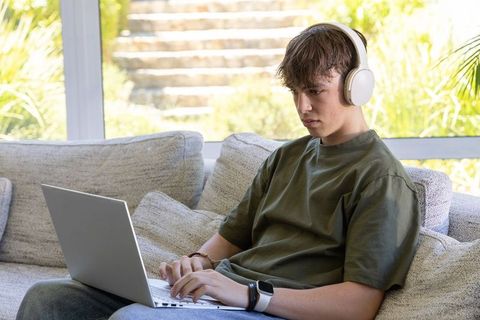 Teen male studying on laptop with wireless headphones on modern sofa by window
