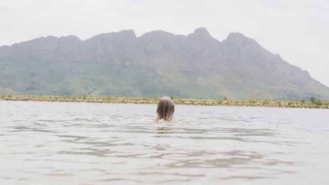 A woman is enjoying her time swimming in a large tranquil lake, surrounded by tall mountains. She wears a bathing suit and seems to be traveling or on vacation. This video could be used to illustrate adventure travel, relaxation, summer vacations, or outdoor activities. It portrays serenity, the beauty of nature, and the joy of outdoor exploration.