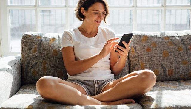 Woman relaxing on sunlit sofa scrolling smartphone in casual loungewear at home