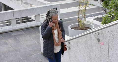 Diverse senior woman walking urban stairs while talking on smartphone and holding railing