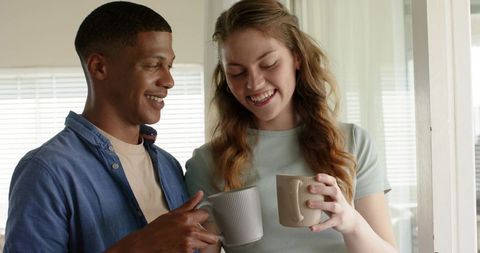 Smiling diverse couple holding ribbed ceramic mugs by sunlit window cozy morning moment