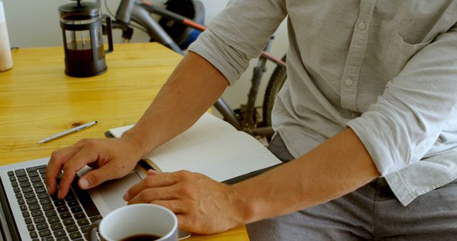A person is working on a laptop while taking notes in an open notebook on a wooden desk. A coffee cup, a pen, and a French press are on the desk. Suitable for projects related to work-from-home setups, productivity, office environments, remote work, and freelance jobs.