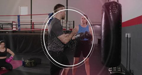Male trainer holding focus mitts coaching female boxer punching heavy bag padwork in gym