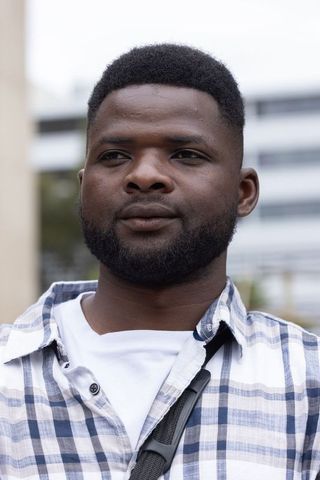 African man standing in urban courtyard looking right wearing plaid shirt and strap