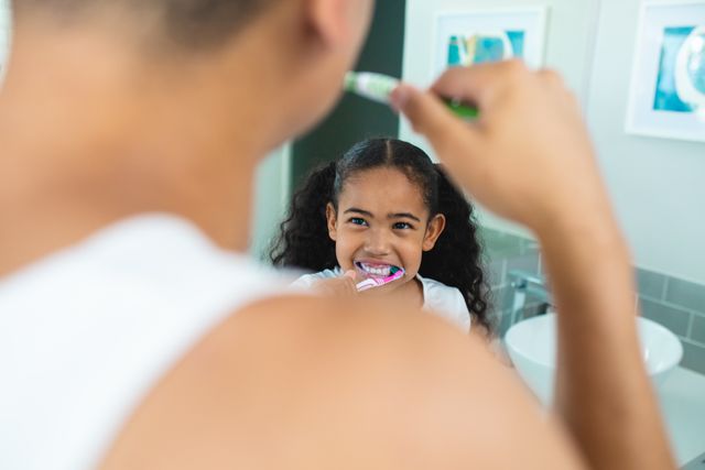 Hispanic girl brushing teeth while looking at father in bathroom at home. unaltered, hygiene, routine, family, lifestyle and togetherness concept.