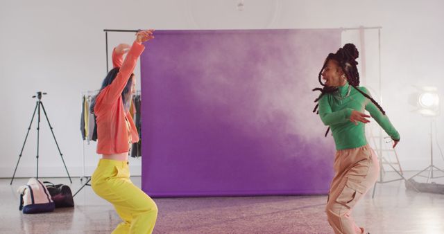 Two women dancing energetically in a studio, dressed in vibrant outfits and standing in front of a purple backdrop. Ideal for use in content related to dance, fitness, wellness, youth culture, and artistic expression.