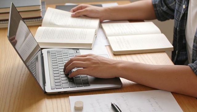 Typing and researching on laptop with open books and handwritten notes on wooden desk