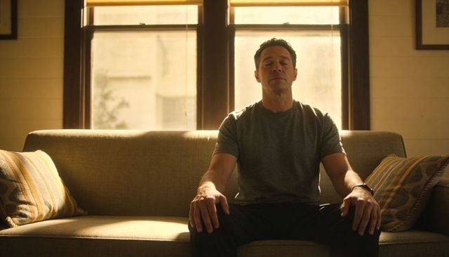 Middle-aged man sitting on sofa meditating at golden hour backlit window calm living room