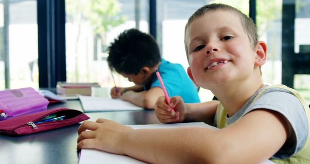 Young boy smiling at camera while doing schoolwork alongside a classmate in a bright classroom. Suitable for educational materials, back-to-school campaigns, and content focused on early childhood education and learning environments.