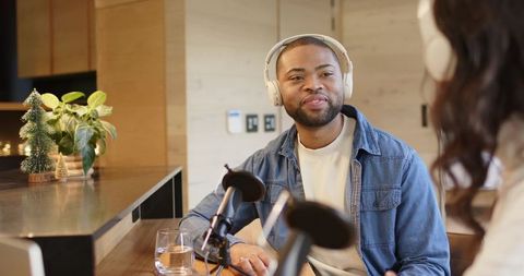 Co-hosts recording relaxed podcast in warm wood cafe, man in denim wearing headphones