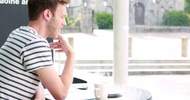 Young man sitting in a modern cafe contemplating by the window. He is wearing a striped shirt and looking thoughtful. Coffee cup on the table adds to the casual ambiance. Perfect for use in content related to thinking, relaxation, cafes, or younger demographics. Great for blogs, lifestyle articles, or advertisements targeting dynamic lifestyle or urban settings.