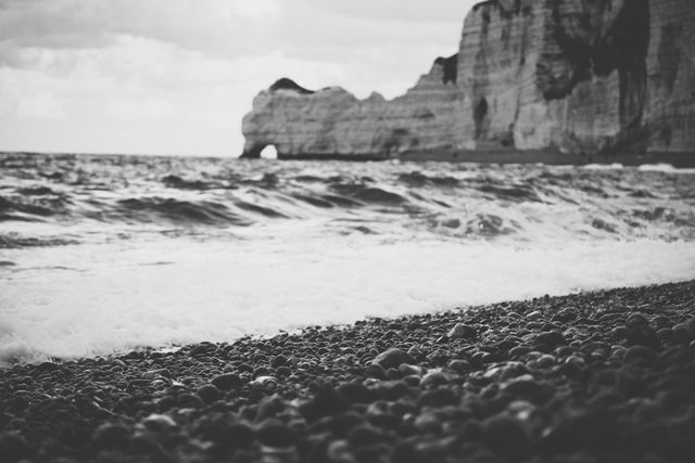 Beautiful black and white depiction of a pebble beach with waves gently crashing onto the shore. The background features stately cliffs rising above the sea, creating a stark contrast with the smooth round pebbles in the foreground. This image is perfect for use in travel blogs, nature-themed websites, or as a calming background in presentations and desktop wallpapers.