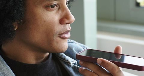 Mid-adult man speaking into smartphone for voice messaging and hands-free communication by window