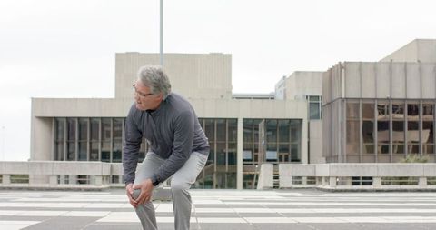 Senior man clutching knee on urban plaza after walking, showing joint pain and instability