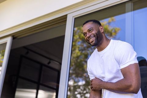 Smiling african man leaning on sliding glass door wearing white t-shirt, modern patio lifestyle