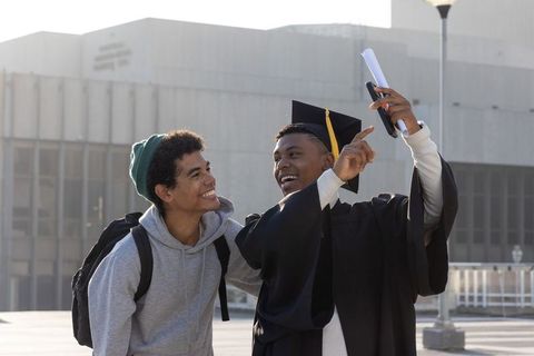 Graduating student taking celebratory selfie with friend in cap and gown holding diploma