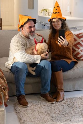 Couple wearing pumpkin hats taking selfie while holding puppy in devil horn headbands