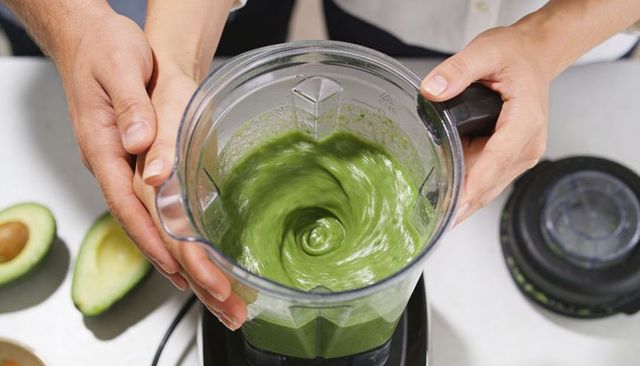 Hands holding blender jar with creamy avocado puree swirling for homemade green smoothie
