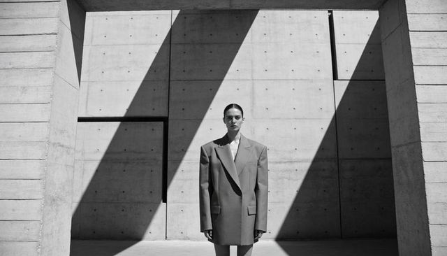 Woman Standing in Oversized Suit in Brutalist Concrete Atrium Highlighted by Diagonal Light