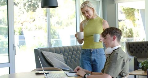 Woman sharing coffee as teen works on laptop with headphones in bright modern home lounge