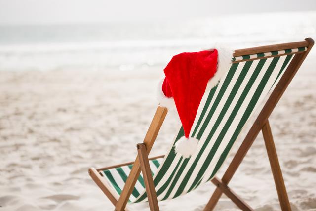Santa hat resting on a green and white striped beach chair at a tropical sand beach. Perfect for holiday-themed travel promotions, Christmas vacation advertisements, and festive greeting cards.